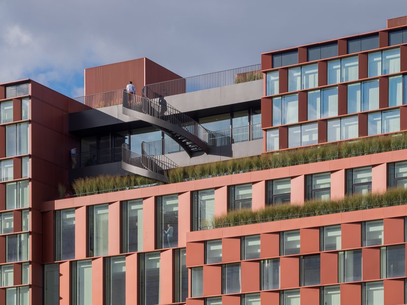 Exterior staircase with planted terraces connecting the red paneled facade levels under cloudy skies