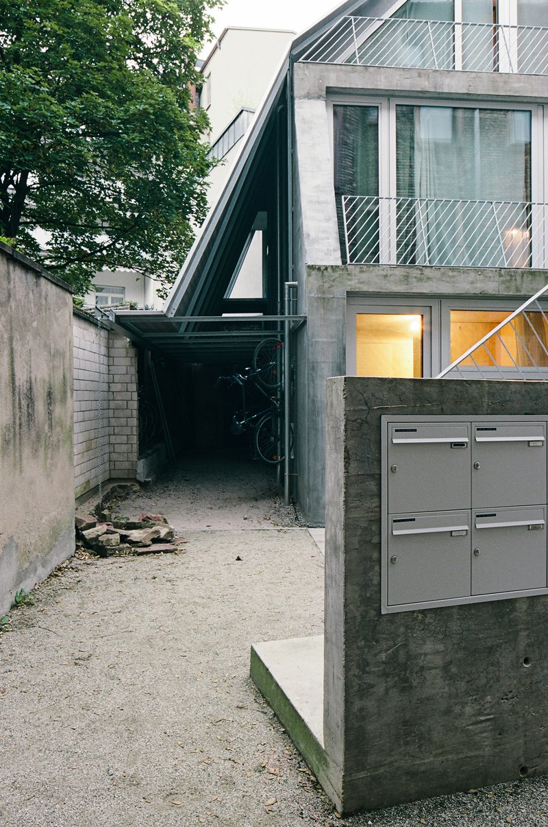 Gravel courtyard entry with black steel frame passageway and concrete mailbox beneath overhanging tree canopy