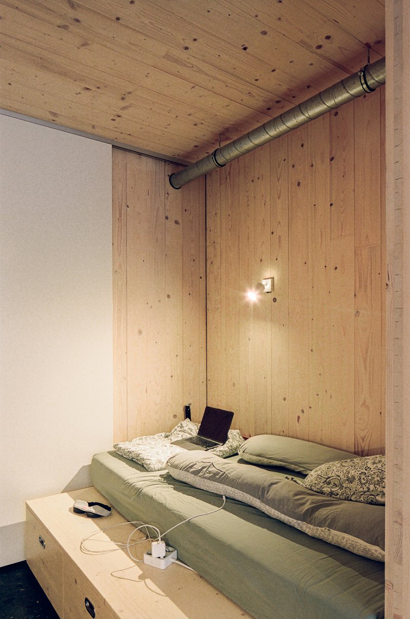 Sleeping platform with raised plywood bed base and exposed ventilation duct along the wood-clad wall