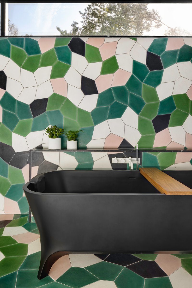 Black freestanding bathtub with timber shelf against geometric tile wall in green, black, and cream tones
