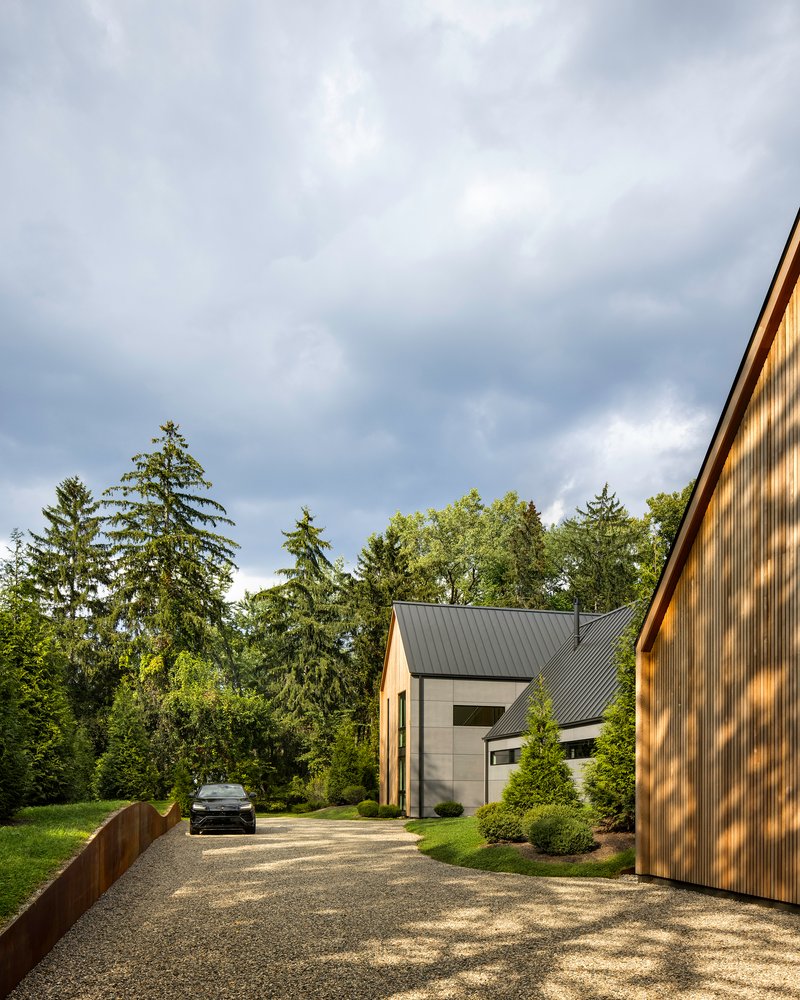 Gravel driveway approach showing white and timber volumes with standing-seam metal roofs under overcast skies
