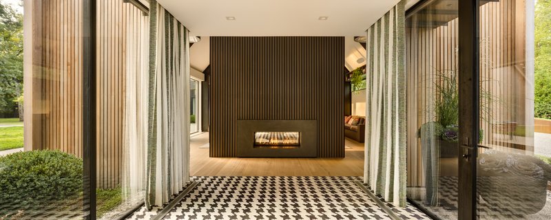 View through glass pavilion with sheer curtains toward the timber-clad fireplace wall and lawn beyond