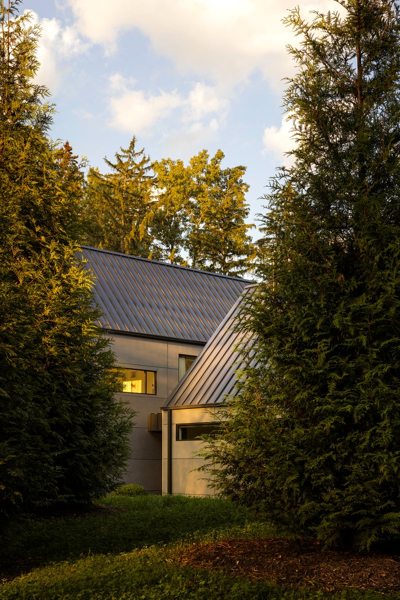 Entry facade with glazed corner and steeply pitched metal roof framed by evergreens at twilight