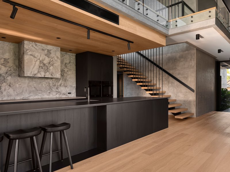 Double-height interior showing floating timber staircase with vertical steel balusters against exposed concrete walls