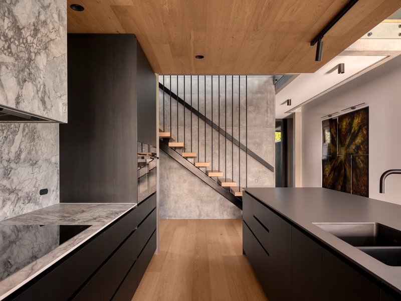 Kitchen with black cabinetry and veined marble countertop beneath timber-clad ceiling and open staircase