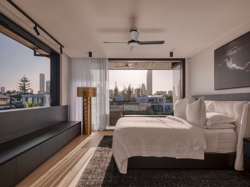 Bedroom with corner glazing and balcony doors framing views of residential rooftops at twilight