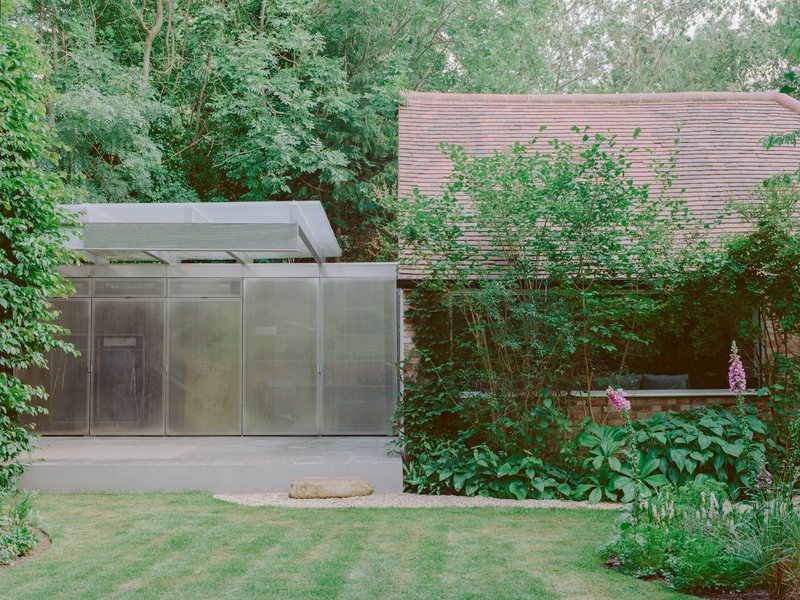 Translucent metal pavilion with flat overhang beside a horizontal timber slatted wall surrounded by garden planting
