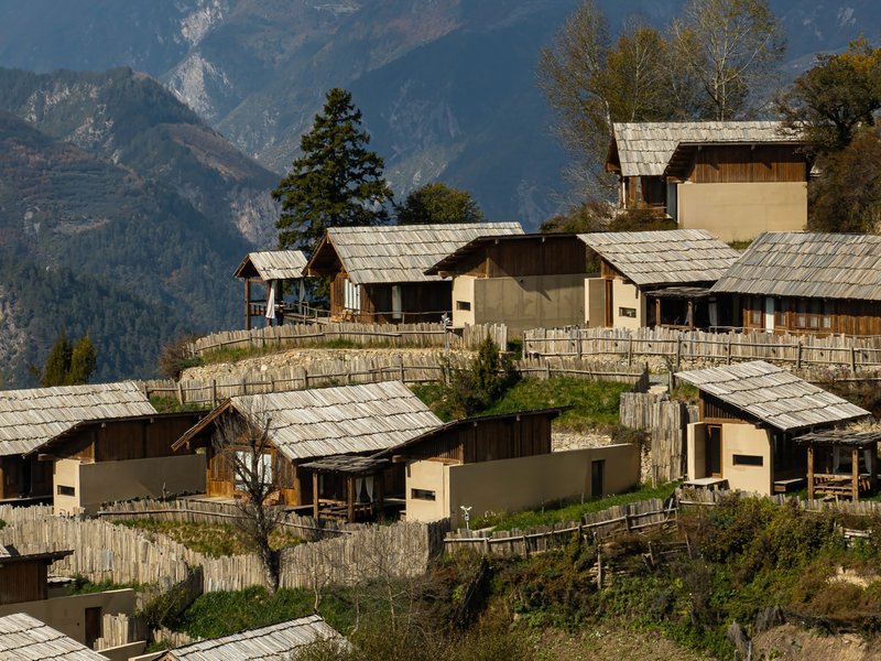 Cluster of timber and stone dwellings with pitched shingled roofs ascending a terraced hillside beneath distant mountains