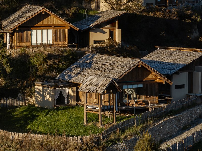Cluster of timber and stone-clad volumes with corrugated metal roofs on a sloping green hillside
