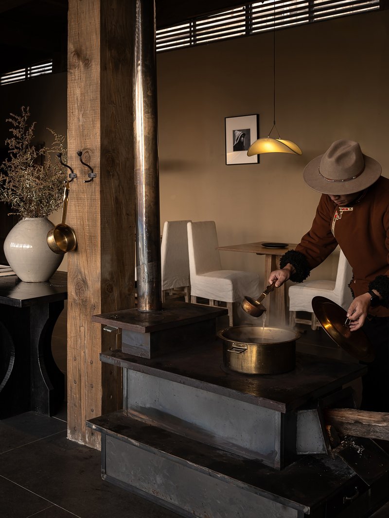 Guest in a wide-brimmed hat pouring tea beside a wood-burning stove under warm pendant light