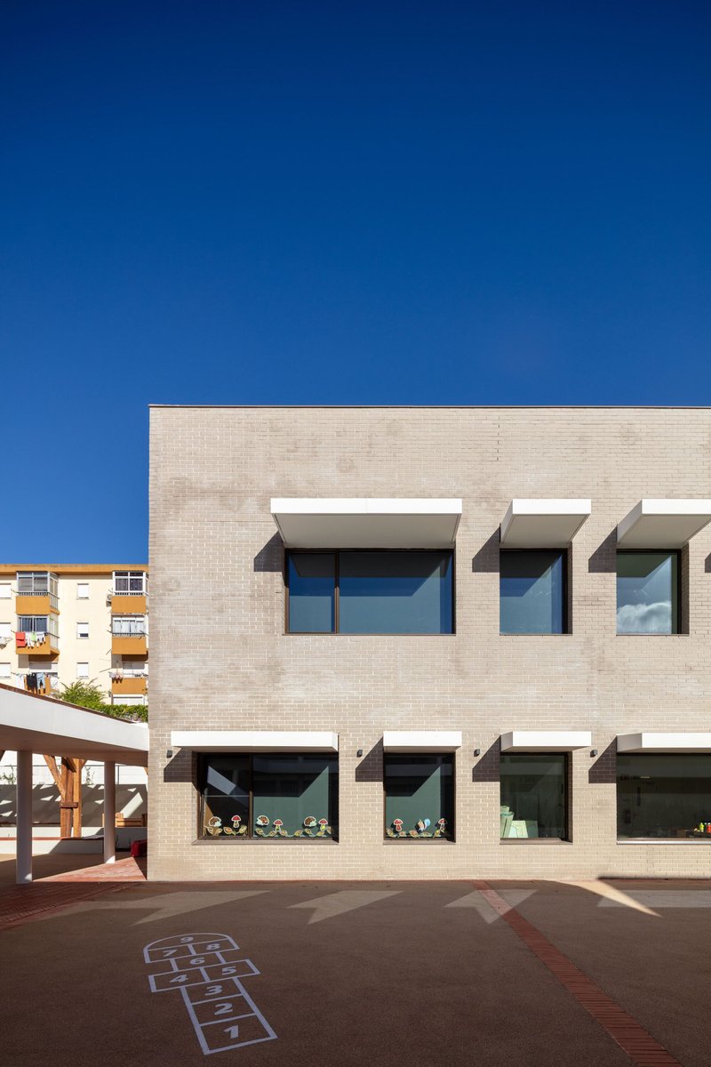 Two-storey buff brick facade with ribbon windows and white canopies overlooking a paved courtyard with hopscotch