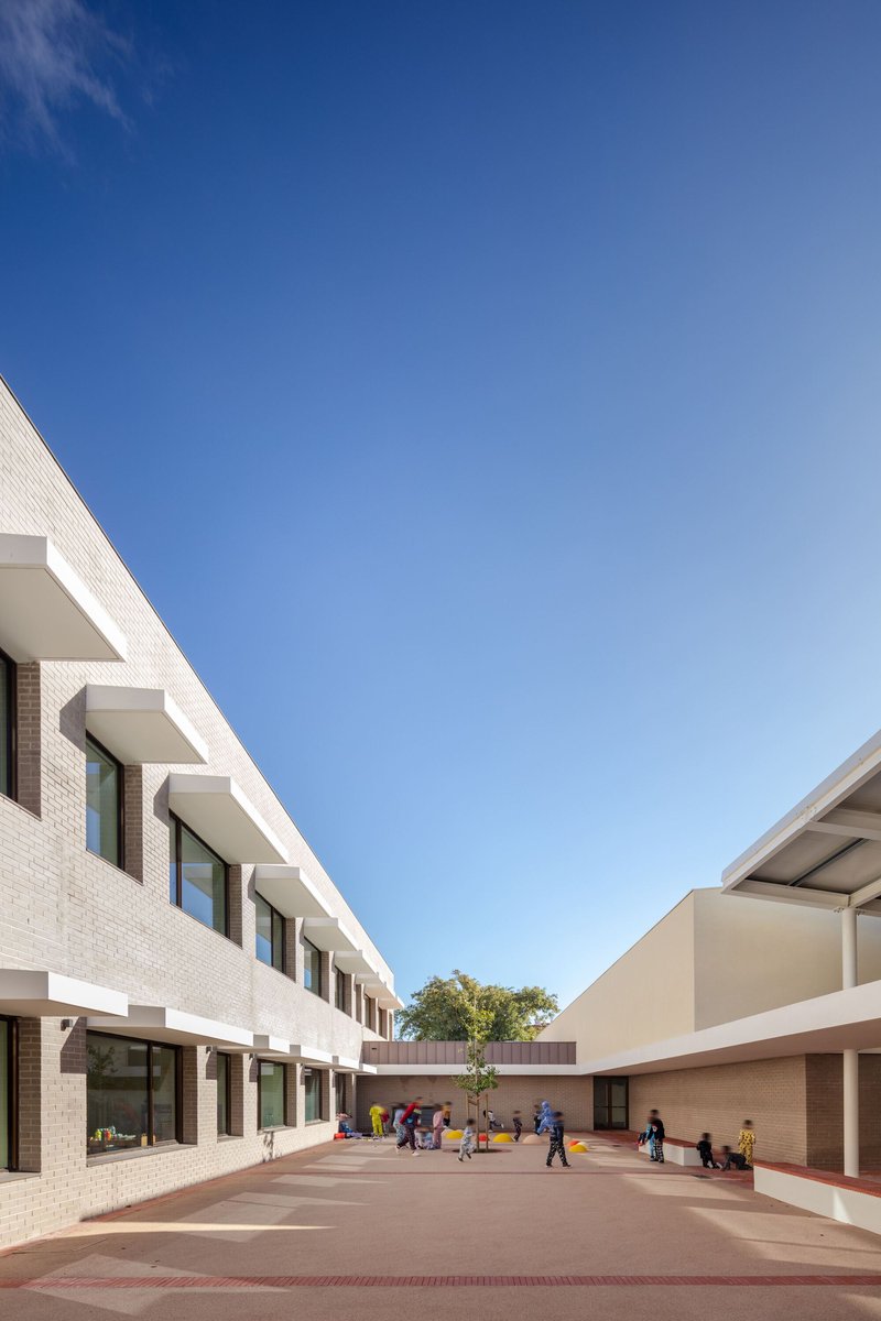 Courtyard view between two brick-clad school wings with children playing on red pavement under a clear blue sky