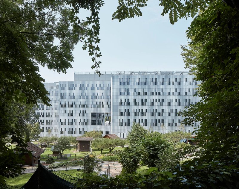 White facade with vertical window pattern framed by overhanging tree branches and a garden plaza below