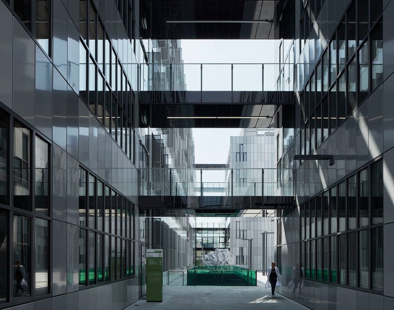 Glazed courtyard with black steel bridges connecting towers as a lone figure walks through sunlit space