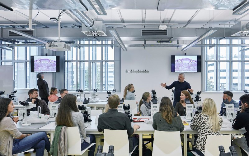 Teaching laboratory with microscopes arranged on white benches beneath exposed ductwork and suspended monitors
