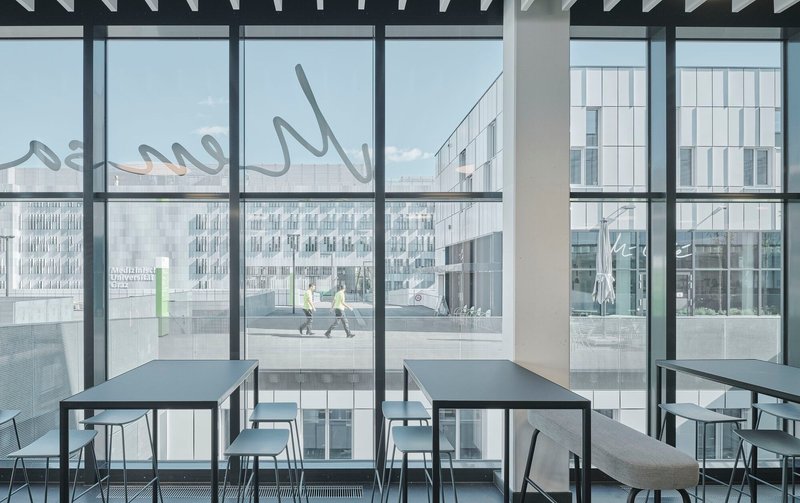Interior dining area with floor-to-ceiling glazing overlooking a plaza where two people walk past