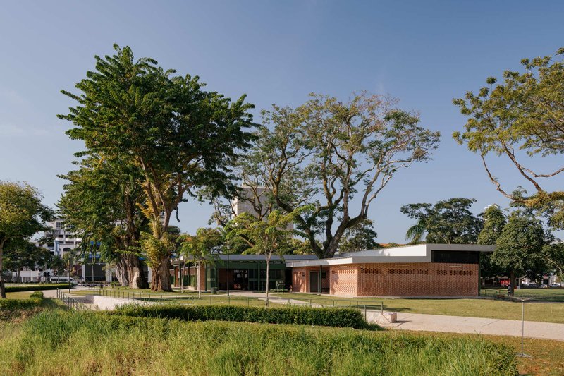 Low-slung brick pavilion with flat roof and clerestory windows set within a landscaped park beneath large trees