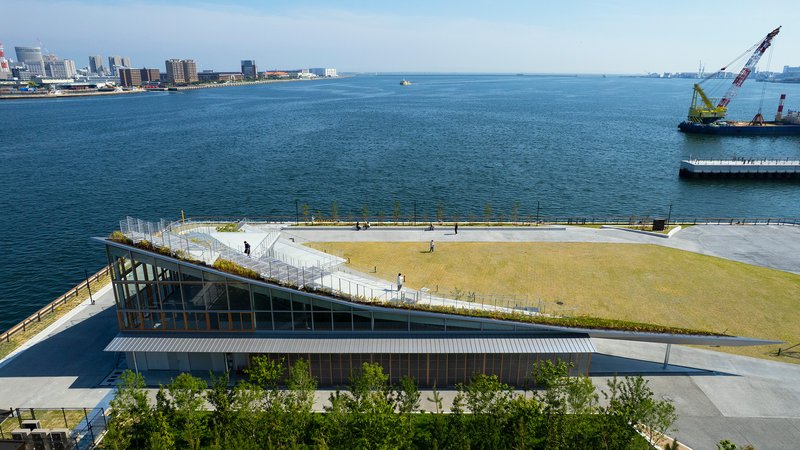 Glass pavilion beneath sloped lawn roof with pedestrians walking on the waterfront promenade