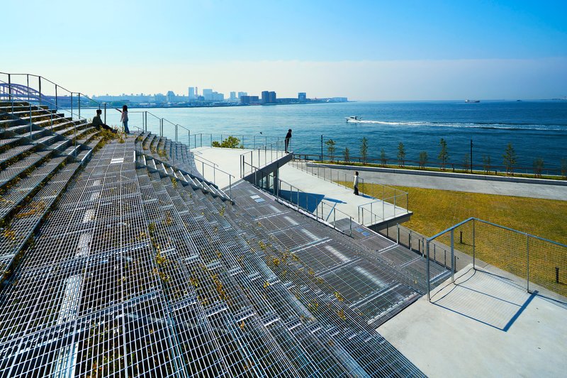 Tiered metal grate seating steps leading down to a waterfront lawn with visitors walking