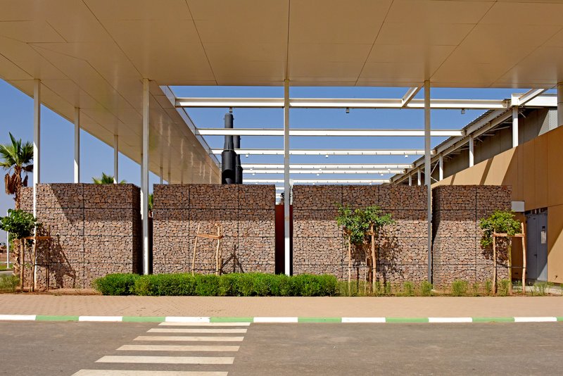 Gabion wall entry with white structural frame overhead and pedestrian crosswalk in front