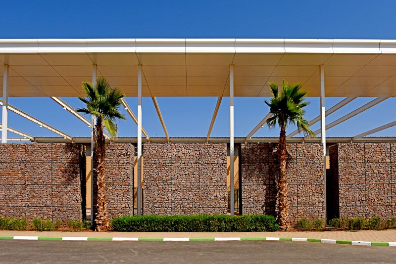 Covered walkway with white columns and diagonal timber bracing behind a gabion wall with palm trees
