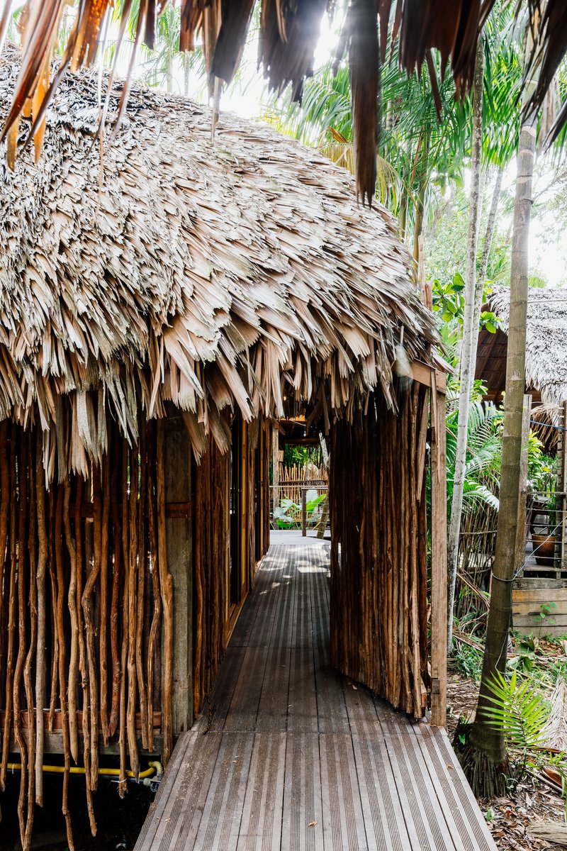 Passageway entrance framed by bundled bamboo walls and layered palm thatch roofing with tropical plantings visible beyond