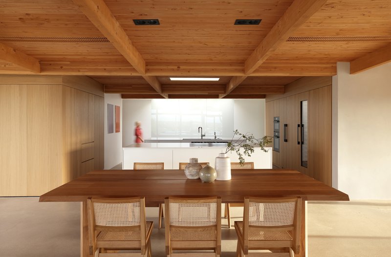 Dining area with timber table and caned chairs beneath heavy timber beams with person moving in kitchen beyond
