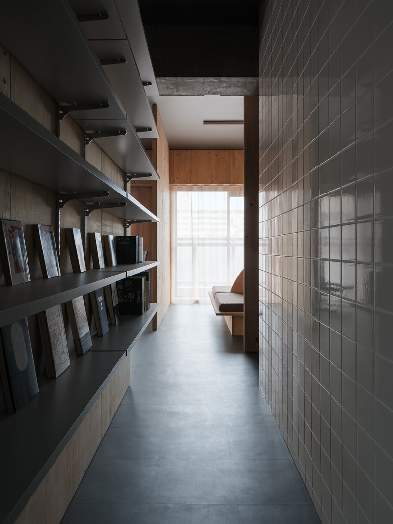 Narrow corridor with black steel shelving and grey tiled wall leading toward a daylit timber-framed opening