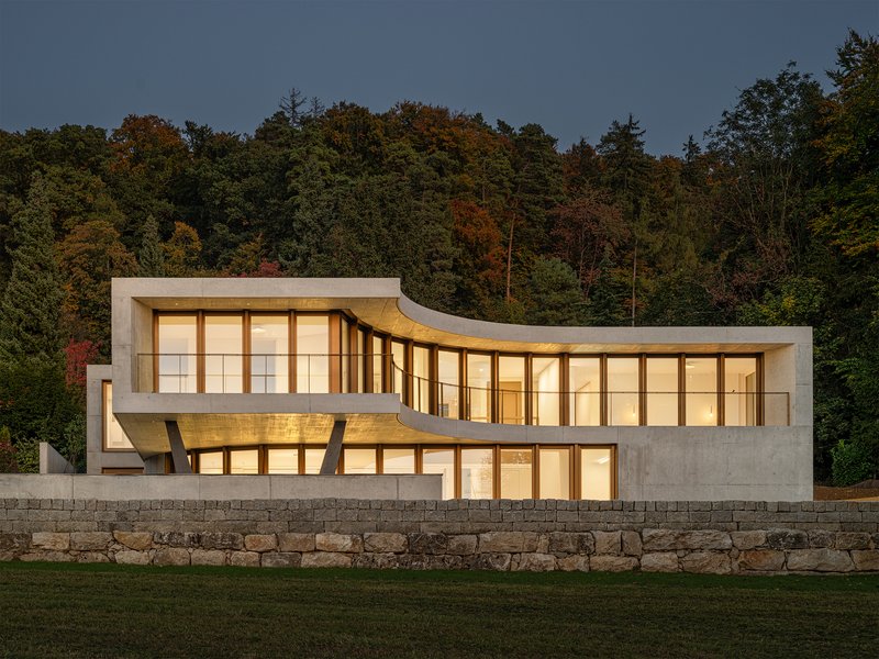 Concrete and timber facade with undulating roofline illuminated at dusk behind stone retaining wall