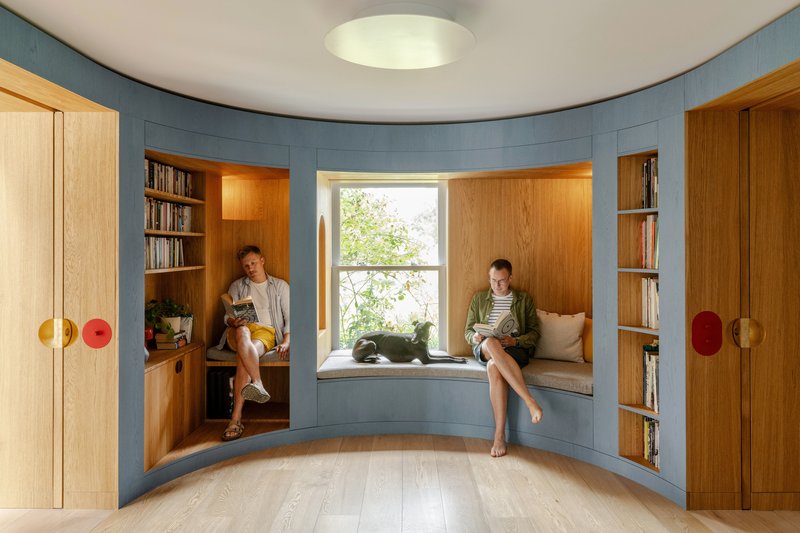 Curved reading room with blue paneling, window seat nook, and two people using built-in shelving alcoves
