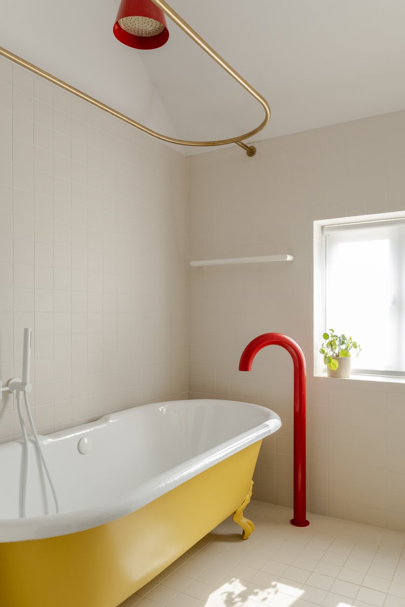 Freestanding tub with yellow base and red floor-mounted tap beneath brass shower rail
