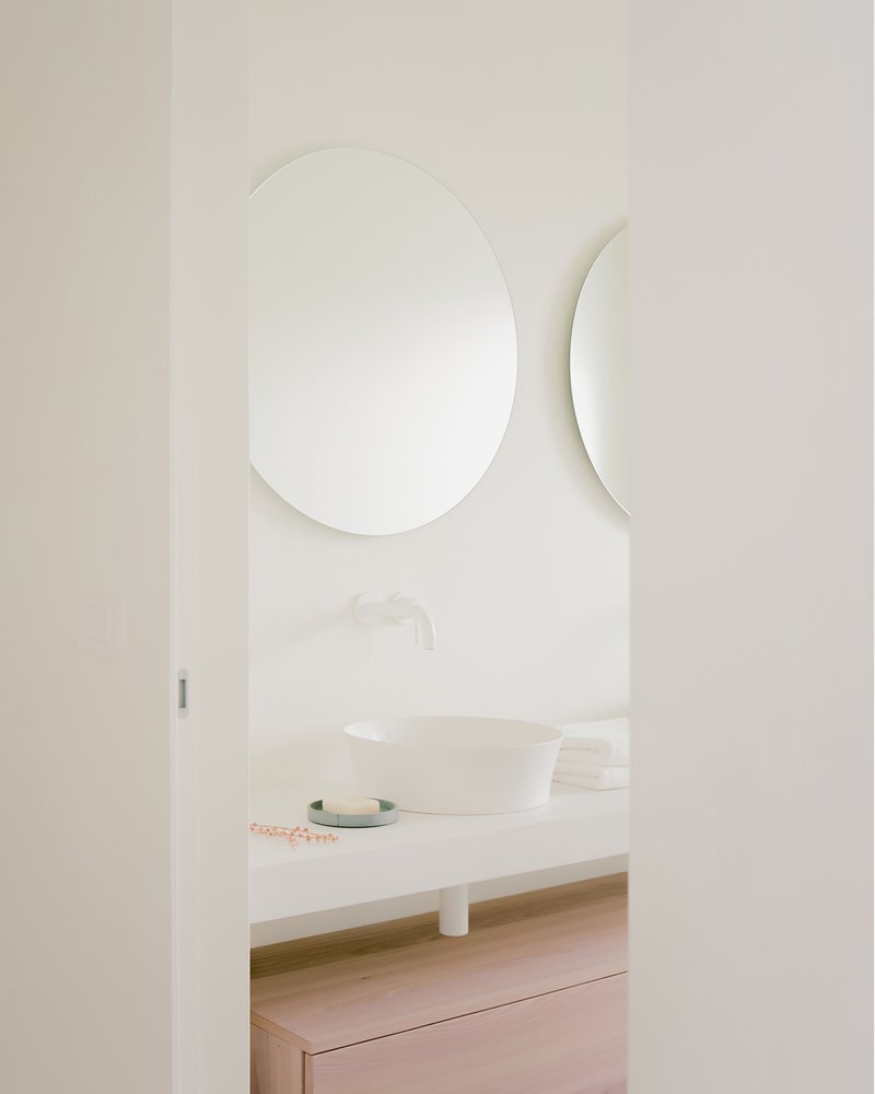 Oval mirror and curved wall-mounted shelf above a vessel sink on pale counter