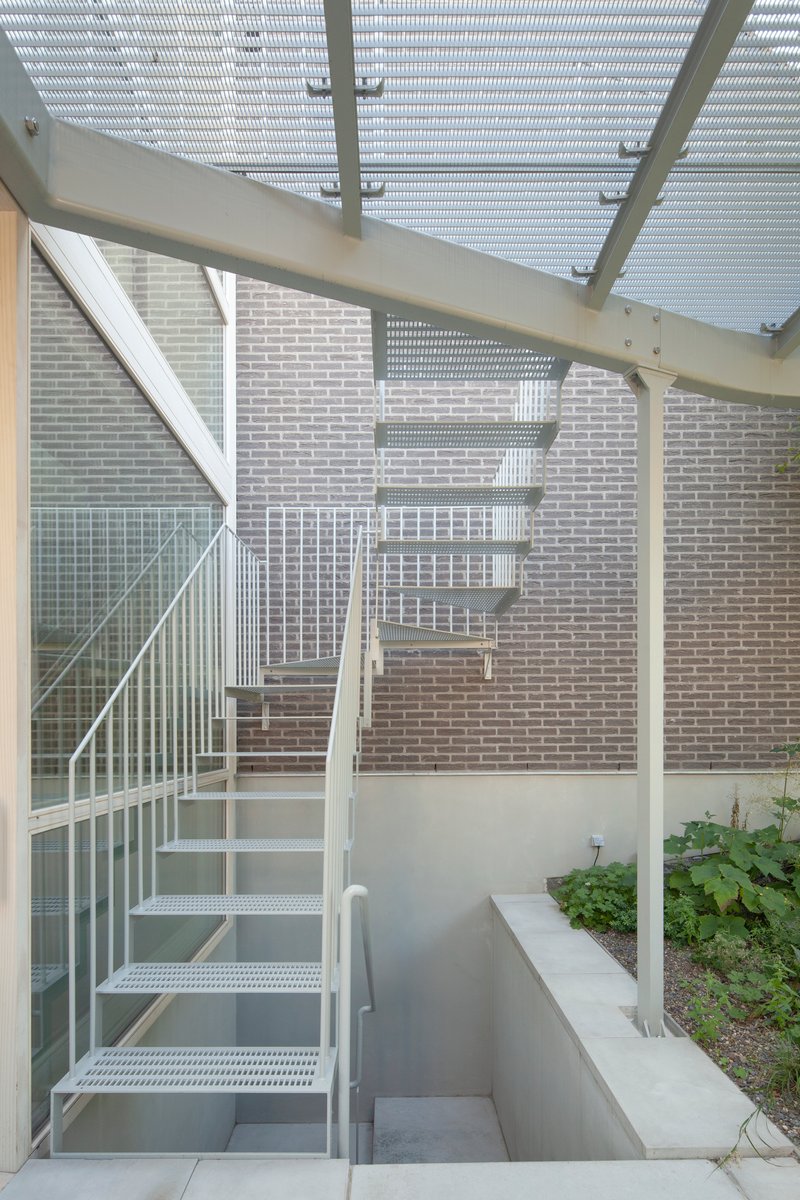 White perforated metal stair with railing ascending alongside a pale brick wall under a slatted pergola