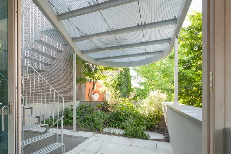 Covered courtyard with translucent corrugated canopy and white metal staircase beside planted bed