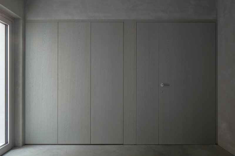 Flush pale grey paneled wall with concealed door and minimal hardware beside a window
