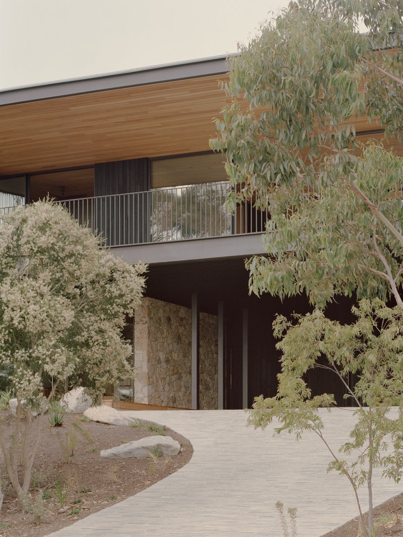 Entry facade with stone-clad walls and timber soffit framed by native plantings and curved pathway