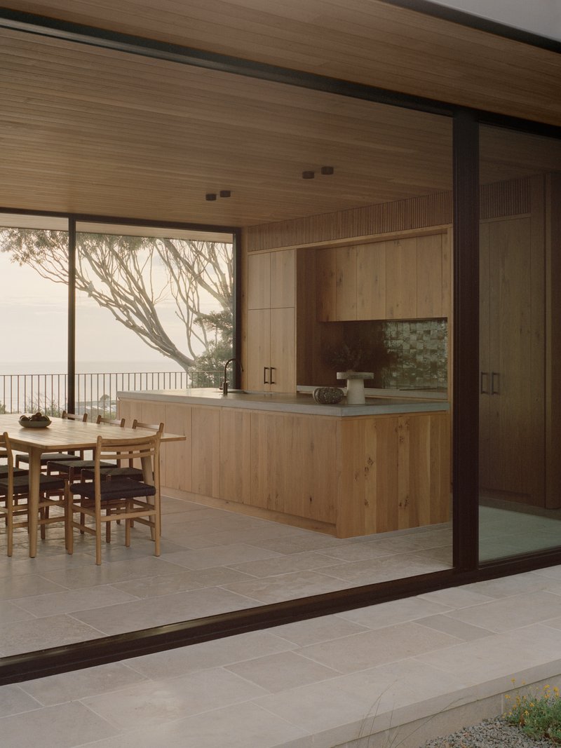 Kitchen island and dining table beneath timber ceiling with ocean view framed by bare tree branches