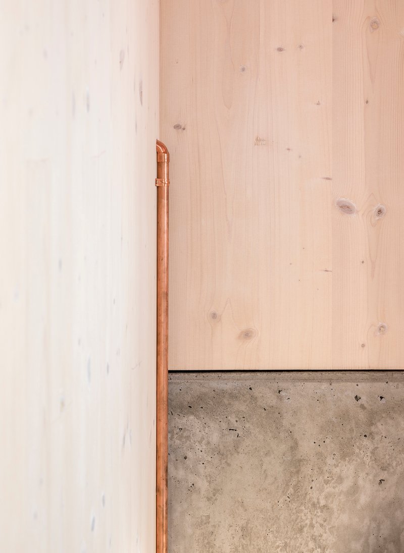 Copper pipe running vertically along white wall above poured concrete plinth and wood panels