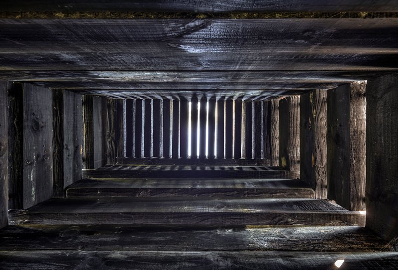 Looking upward through a timber stairwell with charred wood treads and vertical posts framing vertical light slots