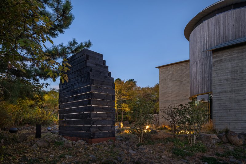 Charred timber tower with horizontal light strips at dusk beside cylindrical concrete volume and planted landscape
