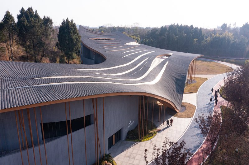 Aerial view of the ribbed metal roof curving between forested hills with visitors on the pathway below