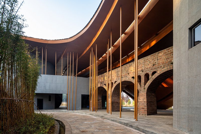 Covered courtyard with vertical timber columns beneath the curving roof and arched brick arcade beyond