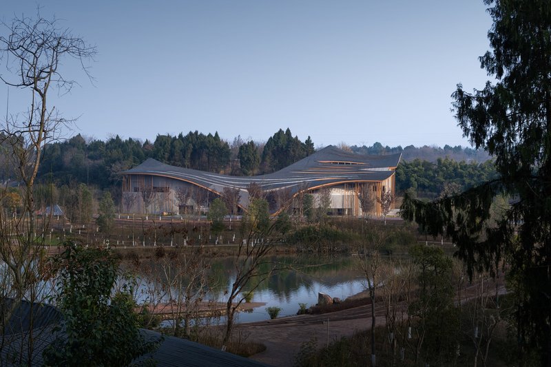 Distant view of the wavelike roof set beside a reflective pond and forested hillside