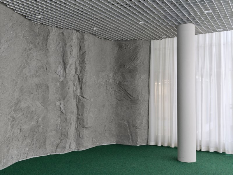 Corner with grey stone wall meeting white sheer curtain, green carpet flooring, and open grid metal ceiling