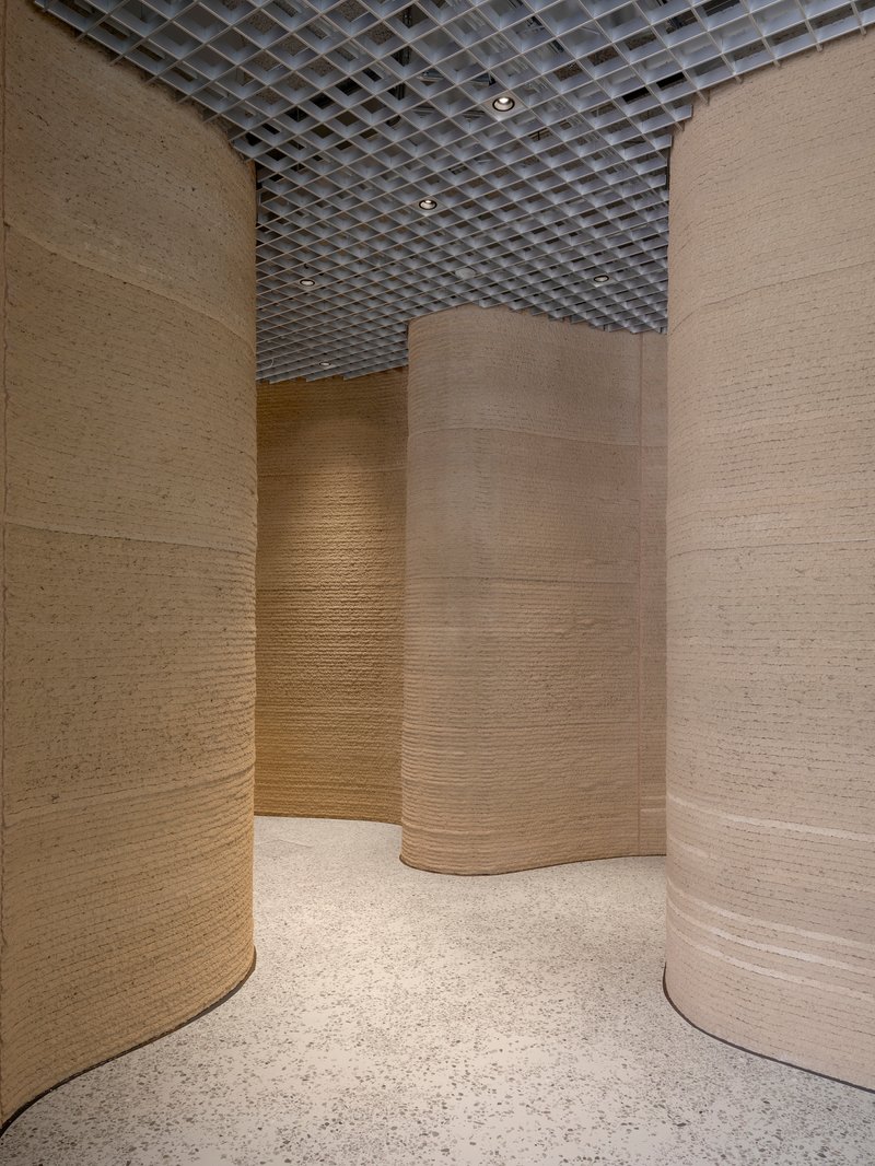 Passage between three curved copper-paneled columns with terrazzo flooring and open grid ceiling above