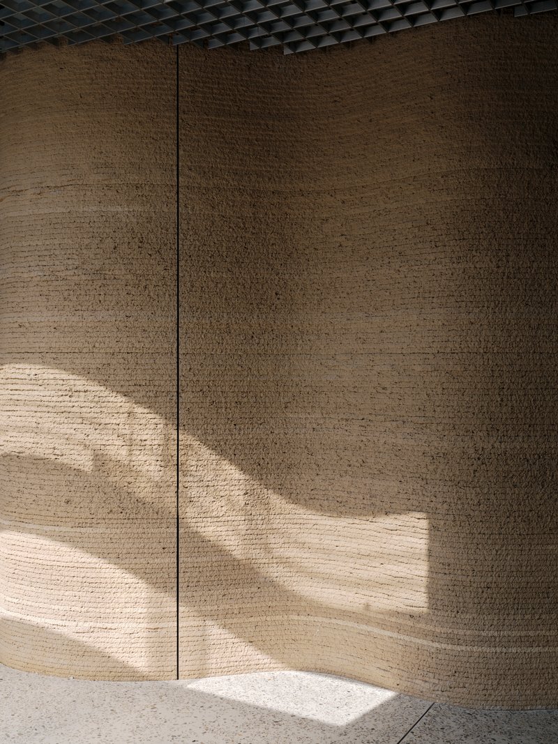 Curved rammed earth wall panels catching diagonal sunlight beneath perforated metal ceiling grid
