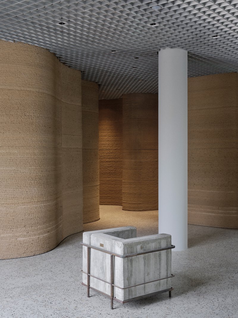 White cylindrical column beside concrete seating cube amid curved timber wall panels and open mesh ceiling