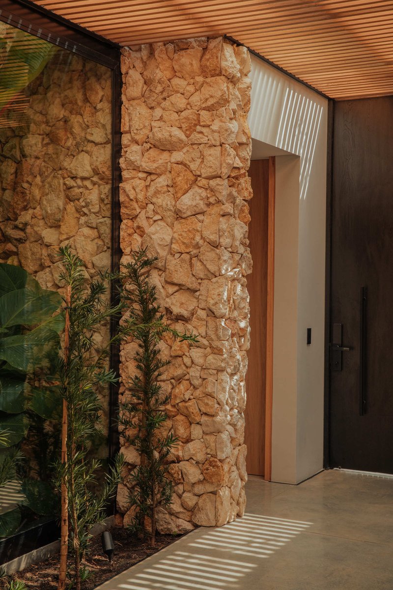 Rough stone wall beside entry door with planted bed and slatted timber ceiling casting linear shadows