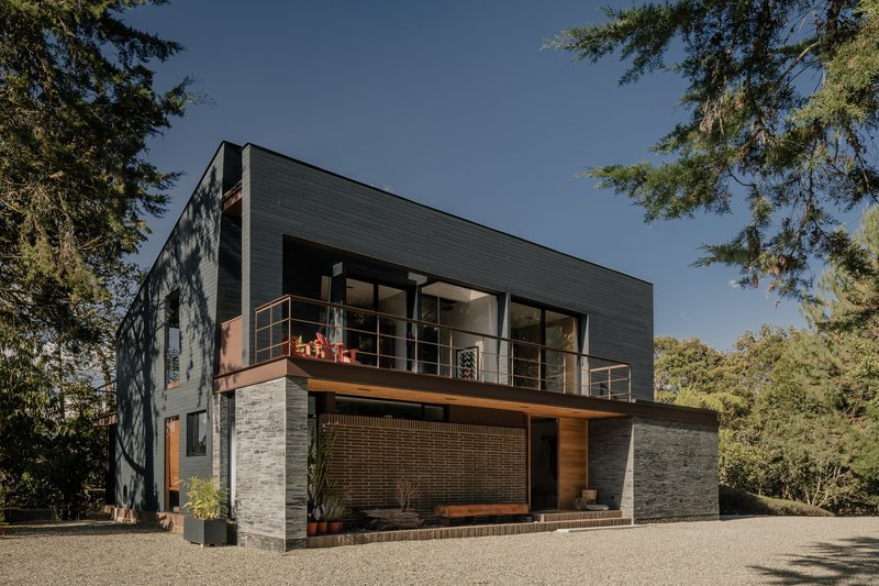 Street view of the two-level volume with metal siding, stone walls and framed balcony between pines