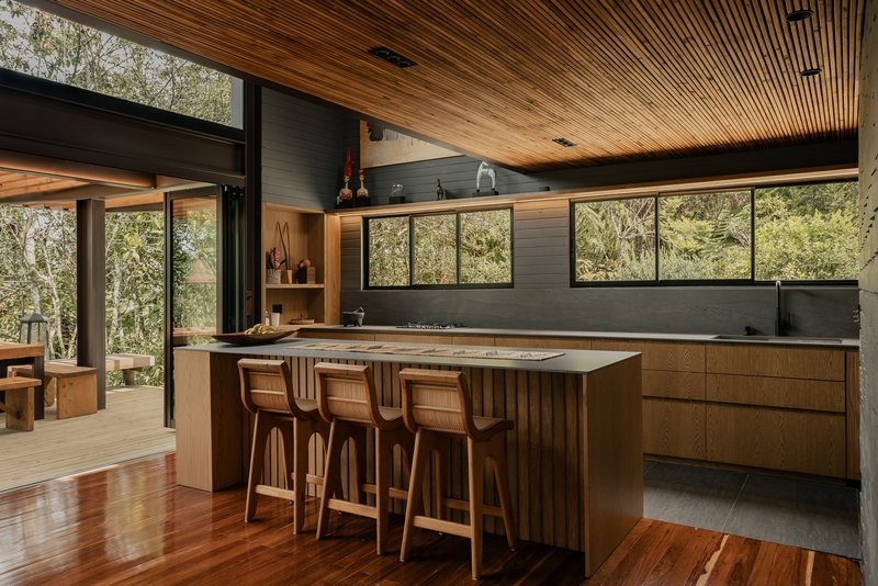 Kitchen island with timber cabinetry and slatted ceiling overlooking forested hillside through clerestory glazing